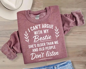 Può includere: Una felpa girocollo bordeaux con la scritta bianca "I CAN'T ARGUE WITH MY Bestie SHE'S OLDER THAN ME AND OLD PEOPLE Don't listen". La felpa è piegata con le maniche arrotolate. Un cappello bianco e jeans blu sono parzialmente visibili.
