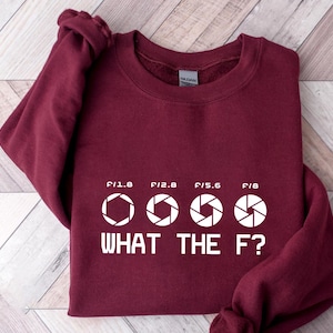 May include: A maroon sweatshirt with white text and graphics. The text reads "WHAT THE F?" below four aperture symbols and the numbers F/1.8, F/2.8, F/5.6, and F/8. The sweatshirt is a casual garment.