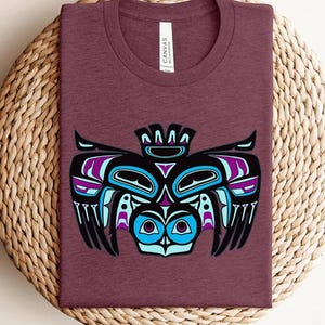 May include: A maroon crew neck t-shirt featuring a graphic print of a stylized bird in black, bright blue, and purple. The design incorporates bold lines and geometric shapes typical of Northwest Coast art. The shirt has a white inner neck label.