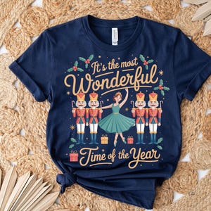 May include: Navy blue t-shirt with a festive design. The shirt features the text "It's the most Wonderful Time of the Year" with illustrations of nutcrackers, a ballerina, and gift boxes. The design is in shades of red, gold, green, and white.