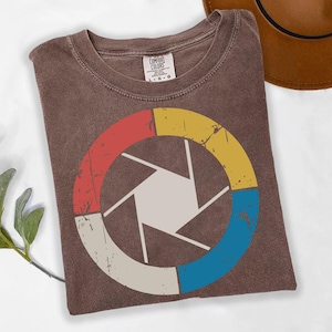 Może przedstawiać: Brązowa koszulka z grafiką przysłony aparatu. Przysłona podzielona na czerwone, żółte, niebieskie i kremowe segmenty. Design ma wygląd vintage. Etykieta Comfort Colors.