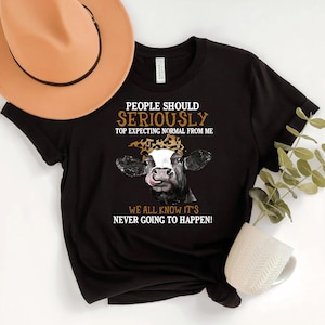 May include: Black t-shirt with a cow graphic wearing a leopard print headband and sticking its tongue out. The text reads "PEOPLE SHOULD SERIOUSLY TOP EXPECTING NORMAL FROM ME WE ALL KNOW IT'S NEVER GOING TO HAPPEN!"