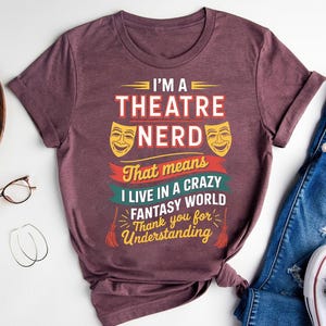 May include: Burgundy t-shirt with the text "I'M A THEATRE NERD" in white and red. The design includes two yellow theater masks and the phrase "That means I LIVE IN A CRAZY FANTASY WORLD". A fun tee for theatre lovers.