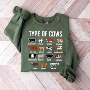 May include: Green sweatshirt with the text "TYPE OF COWS" and illustrations of various cow breeds, including Black Angus, Charolais, Hereford, and Simmental. The sweatshirt is made of a soft material.