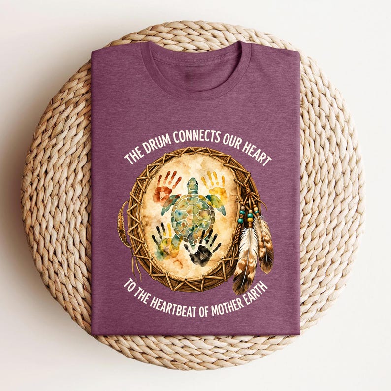 A purple t-shirt with a design featuring a turtle and feathers, along with the text "The drum connects our heart to the heartbeat of mother earth".