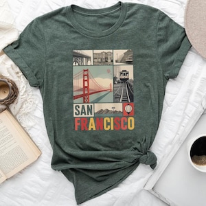 Puede incluir: Una camiseta verde oscuro con un gráfico de los lugares emblemáticos de San Francisco, incluido el puente Golden Gate y un tranvía. El texto "SAN FRANCISCO" está impreso en letras de bloque coloridas debajo de las imágenes. La camiseta es de material suave.