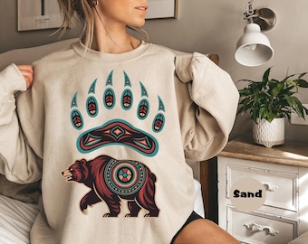 May include: Beige sweatshirt featuring a large bear paw print above a bear illustration. The bear and paw print are in shades of brown, teal, and red. The word "Sand" is printed on a circular label.