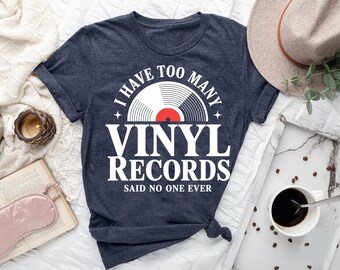 Può includere: T-shirt blu navy con la scritta bianca "I HAVE TOO MANY VINYL RECORDS SAID NO ONE EVER" e un design a vinile. L'immagine include un cappello, caffè e una candela.