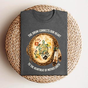 A gray t-shirt with a design featuring a turtle and feathers, along with the text "The drum connects our heart to the heartbeat of mother earth".