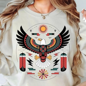 May include: Cream-colored sweatshirt featuring a stylized eagle design with outstretched wings. The eagle is depicted in black, red, green, and yellow, with a white head and chest. The design includes a sun, feathers, and geometric shapes, creating a Native American-inspired aesthetic.