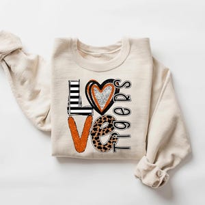 May include: Cream-colored sweatshirt with the word "LOVE" in a graphic design. The "L" is black and white striped, the "O" is a heart with silver glitter, and the "V" is orange. The word "Tigers" is below the word "LOVE".