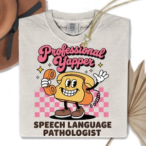 Puede incluir: Camiseta de color beige claro con las palabras "Professional Yapper" en letras rosas y negras, con un personaje de teléfono de dibujos animados. La camiseta también incluye el texto "Speech Language Pathologist" debajo del gráfico.