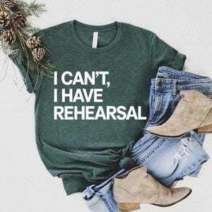 May include: Dark green t-shirt with the text "I CAN'T, I HAVE REHEARSAL" in white. The shirt is paired with blue denim jeans and two pairs of tan ankle boots. A decorative pine branch with pine cones is placed on the top left.