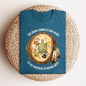 A blue t-shirt with a design featuring a turtle and feathers, along with the text "The drum connects our heart to the heartbeat of mother earth".