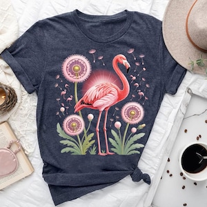 May include: A navy blue t-shirt featuring a pink flamingo design with dandelions. The flamingo is surrounded by pink dandelions and smaller flamingo illustrations. The shirt is tied at the bottom.