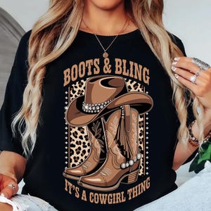 May include: Black t-shirt featuring a graphic of a cowboy hat and boots with the text "BOOTS & BLING" and "IT'S A COWGIRL THING". The design includes brown and tan colors with a leopard print background.