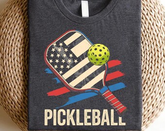Può includere: T-shirt grigio scuro con un design di racchetta da pickleball con la bandiera americana e una palla verde. Strisce rosse e blu sono dietro la racchetta e la parola "PICKLEBALL" è stampata in grassetto.