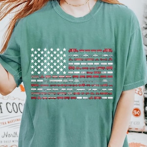 May include: Teal t-shirt with a graphic of the American flag, where the stripes are made of red and white train carriages. The stars are white. A distinctive design on a casual tee.