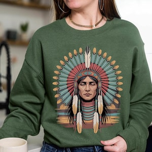 May include: Green sweatshirt featuring a Native American illustration. The graphic depicts a person wearing a headdress with feathers in shades of teal, red, and yellow. The person has their eyes closed. The sweatshirt is a solid green color.