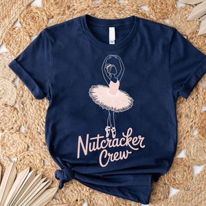 May include: Dark blue t-shirt with a ballerina design in a pink tutu. The words "Nutcracker Crew" are written in a cursive font below the ballerina. The shirt is made of soft material.