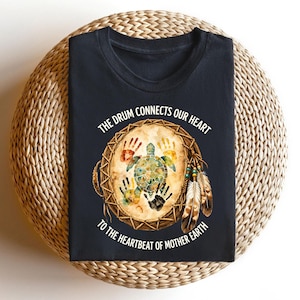A black t-shirt with a design featuring a drum surrounded by feathers and a turtle, along with the text "The drum connects our heart to the heartbeat of mother earth".