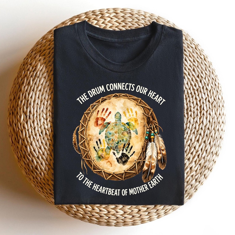 A black t-shirt with a design featuring a drum surrounded by feathers and a turtle, along with the text "The drum connects our heart to the heartbeat of mother earth".