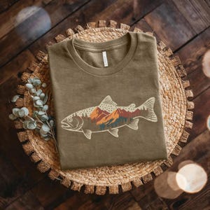 Peut inclure: T-shirt vert olive avec un motif de poisson orné d'un paysage de montagnes et de forêts. L'illustration du poisson utilise des tons orange, marron et vert. Le t-shirt est plié sur un tapis tissé, créant une esthétique inspirée de la nature.