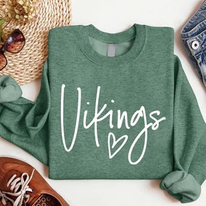 May include: A green crew neck sweatshirt with the word "Vikings" in white script and a small heart. The sweatshirt is folded, with the sleeves rolled up. The image also includes brown shoes, sunglasses, and denim shorts.