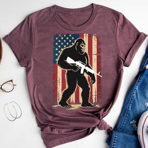 Patriotic Bigfoot Shirt – Armed Sasquatch Graphic Tee – USA Flag Cryptid Shirt – Bold Survivalist Gift – American Outdoorsman T-Shirt