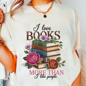 May include: Cream-coloured t-shirt with the text "I love BOOKS MORE THAN I like people." The graphic features a stack of books with floral accents in pink, purple, and brown.