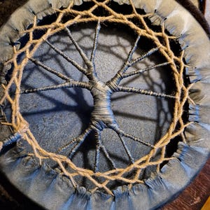 May include: A round, blue leather drum with a tree-like design in the center. The drum is surrounded by a woven rope border.