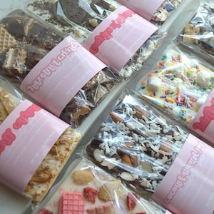 Peut inclure: Vue a&eacute;rienne de plusieurs barres de chocolat, chacune emball&eacute;e individuellement. Les garnitures varient : biscuits, noix et vermicelles. Chaque paquet est entour&eacute; d'une &eacute;tiquette rose avec le texte "Hazelnut & Joy".