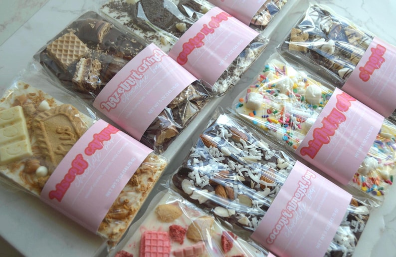 Peut inclure: Vue a&eacute;rienne de plusieurs barres de chocolat, chacune emball&eacute;e individuellement. Les garnitures varient : biscuits, noix et vermicelles. Chaque paquet est entour&eacute; d'une &eacute;tiquette rose avec le texte "Hazelnut & Joy".