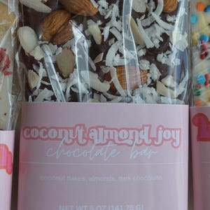 Peut inclure: Une barre de chocolat noir garnie d'amandes et de flocons de noix de coco, emball&eacute;e dans du plastique transparent. L'&eacute;tiquette rose indique "coconut almond joy chocolate bar". Poids net 141,75 g.