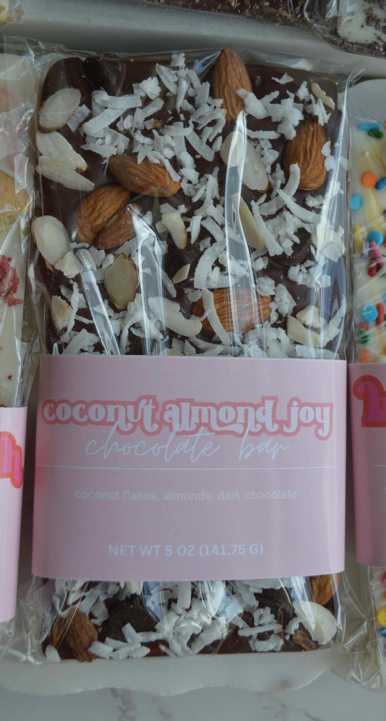 Peut inclure: Une barre de chocolat noir garnie d'amandes et de flocons de noix de coco, emball&eacute;e dans du plastique transparent. L'&eacute;tiquette rose indique "coconut almond joy chocolate bar". Poids net 141,75 g.