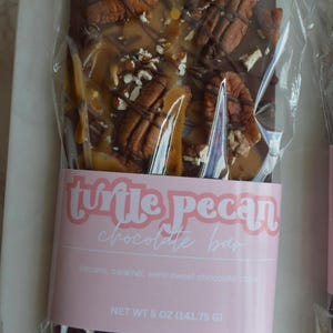 Peut inclure: Une barre de chocolat aux noix de p&eacute;can et au caramel, emball&eacute;e dans du plastique transparent. La barre porte une &eacute;tiquette rose avec l'inscription "turtle pecan chocolate bar". La barre p&egrave;se 141,75 g.