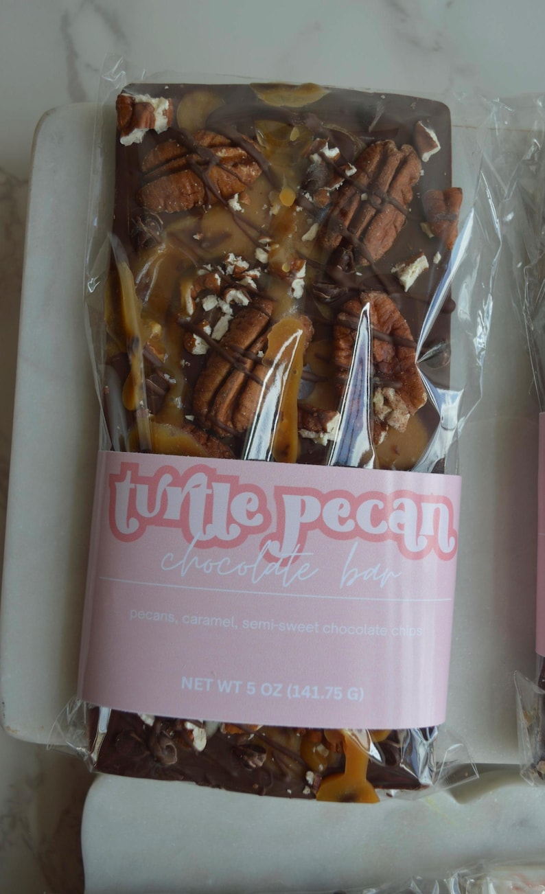 Peut inclure: Une barre de chocolat aux noix de p&eacute;can et au caramel, emball&eacute;e dans du plastique transparent. La barre porte une &eacute;tiquette rose avec l'inscription "turtle pecan chocolate bar". La barre p&egrave;se 141,75 g.