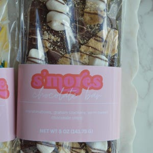 Peut inclure: Une barre de chocolat avec des guimauves, des biscuits Graham et des p&eacute;pites de chocolat mi-sucr&eacute;. La barre est envelopp&eacute;e dans du plastique transparent et porte une &eacute;tiquette rose avec l'inscription "s'mores chocolate bar". Poids net : 141,75 g.