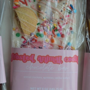 Peut inclure: Une barre de chocolat aux biscuits animaux givr&eacute;s dans un emballage transparent. La barre est en chocolat blanc avec des vermicelles color&eacute;s, un filet rose et des biscuits animaux. L'&eacute;tiquette indique "Frosted, animal, cook chocolate bar". Poids net 141,75 g.