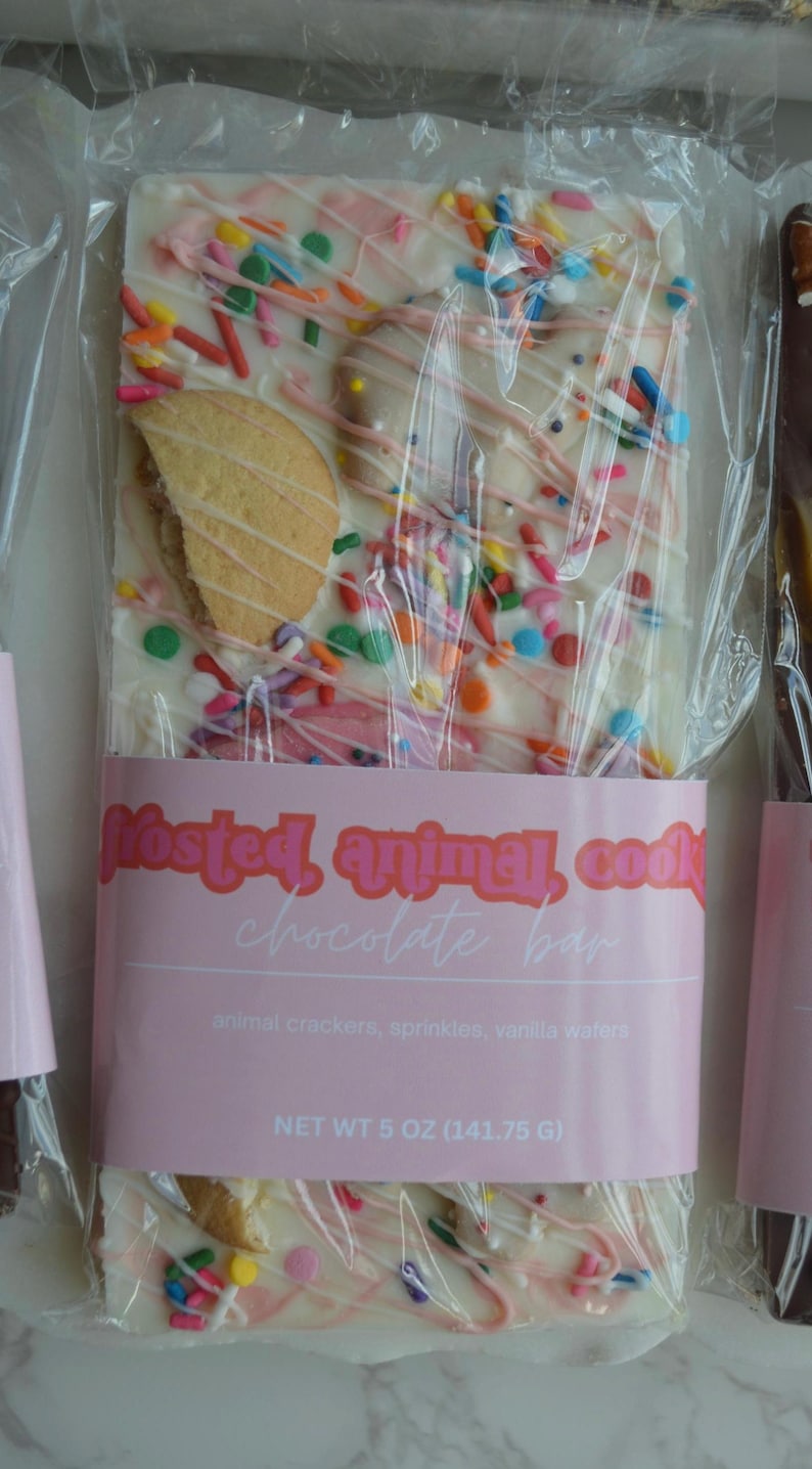 Peut inclure: Une barre de chocolat aux biscuits animaux givr&eacute;s dans un emballage transparent. La barre est en chocolat blanc avec des vermicelles color&eacute;s, un filet rose et des biscuits animaux. L'&eacute;tiquette indique "Frosted, animal, cook chocolate bar". Poids net 141,75 g.