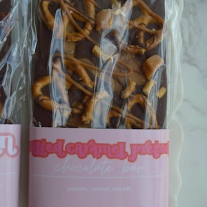 Peut inclure: Une barre de chocolat noir garnie de bretzels et de caramel, emball&eacute;e dans du plastique transparent. L'&eacute;tiquette rose indique "Barre chocolat&eacute;e au caramel sal&eacute; et aux bretzels". Poids net : 141,75 g.
