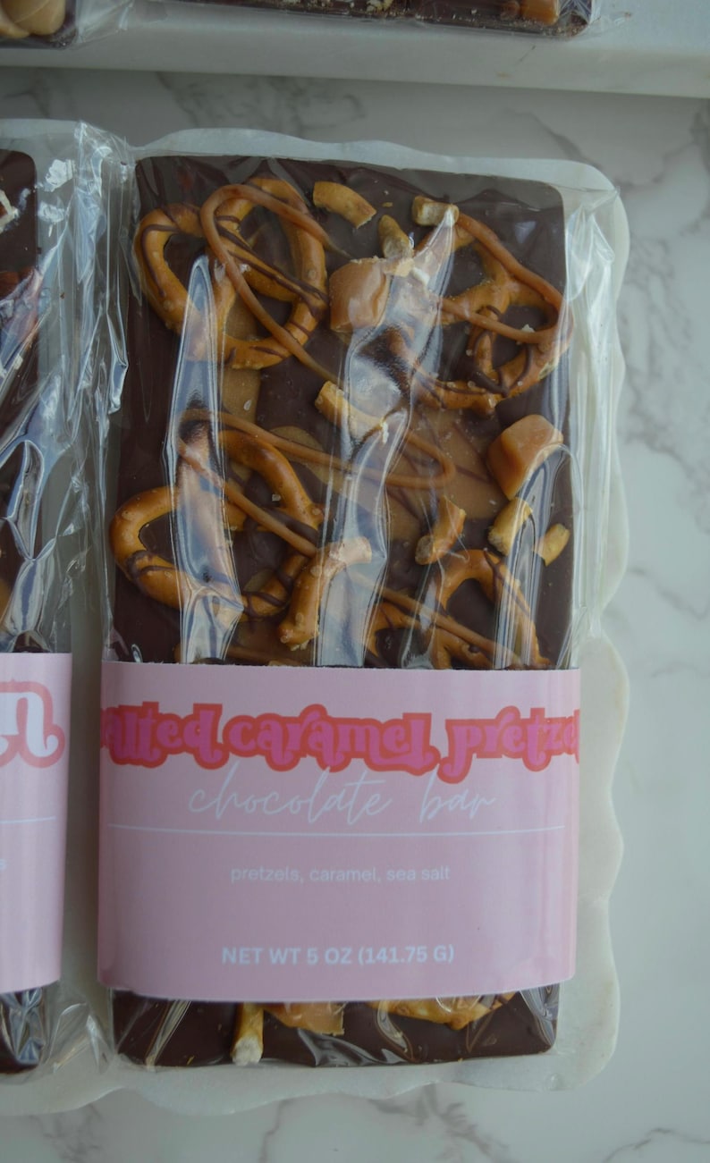 Peut inclure: Une barre de chocolat noir garnie de bretzels et de caramel, emball&eacute;e dans du plastique transparent. L'&eacute;tiquette rose indique "Barre chocolat&eacute;e au caramel sal&eacute; et aux bretzels". Poids net : 141,75 g.
