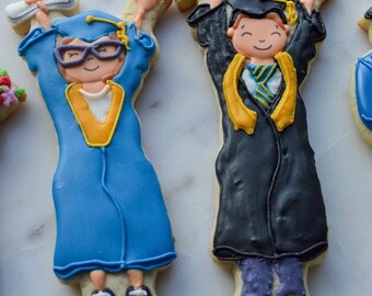Peut inclure: Biscuits sur le thème de la remise des diplômes. Un biscuit bleu avec des lunettes et un col jaune, tenant un diplôme. L'autre biscuit est noir avec une écharpe jaune, tenant un diplôme. Les deux biscuits ont des visages souriants et sont décorés de glaçage.