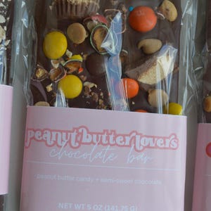 Peut inclure: Une barre de chocolat noir avec des bonbons au beurre de cacahu&egrave;te et du chocolat mi-sucr&eacute;, garnie de divers bonbons. La barre est envelopp&eacute;e dans du plastique transparent avec une &eacute;tiquette rose indiquant "peanut butter lovers chocolate bar".