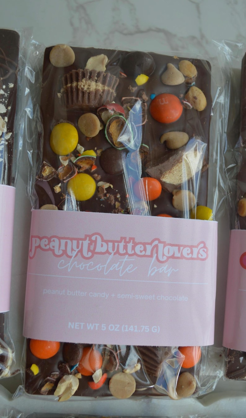 Peut inclure: Une barre de chocolat noir avec des bonbons au beurre de cacahu&egrave;te et du chocolat mi-sucr&eacute;, garnie de divers bonbons. La barre est envelopp&eacute;e dans du plastique transparent avec une &eacute;tiquette rose indiquant "peanut butter lovers chocolate bar".