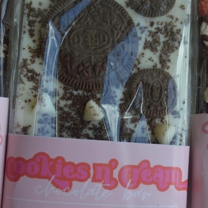 Peut inclure: Une barre de chocolat Cookies n' Cream avec des biscuits Oreo et des p&eacute;pites de chocolat blanc. La barre est envelopp&eacute;e dans du plastique transparent et porte une &eacute;tiquette rose avec le texte "Cookies n' Cream Chocolate Bar". Poids net 141,75 g.