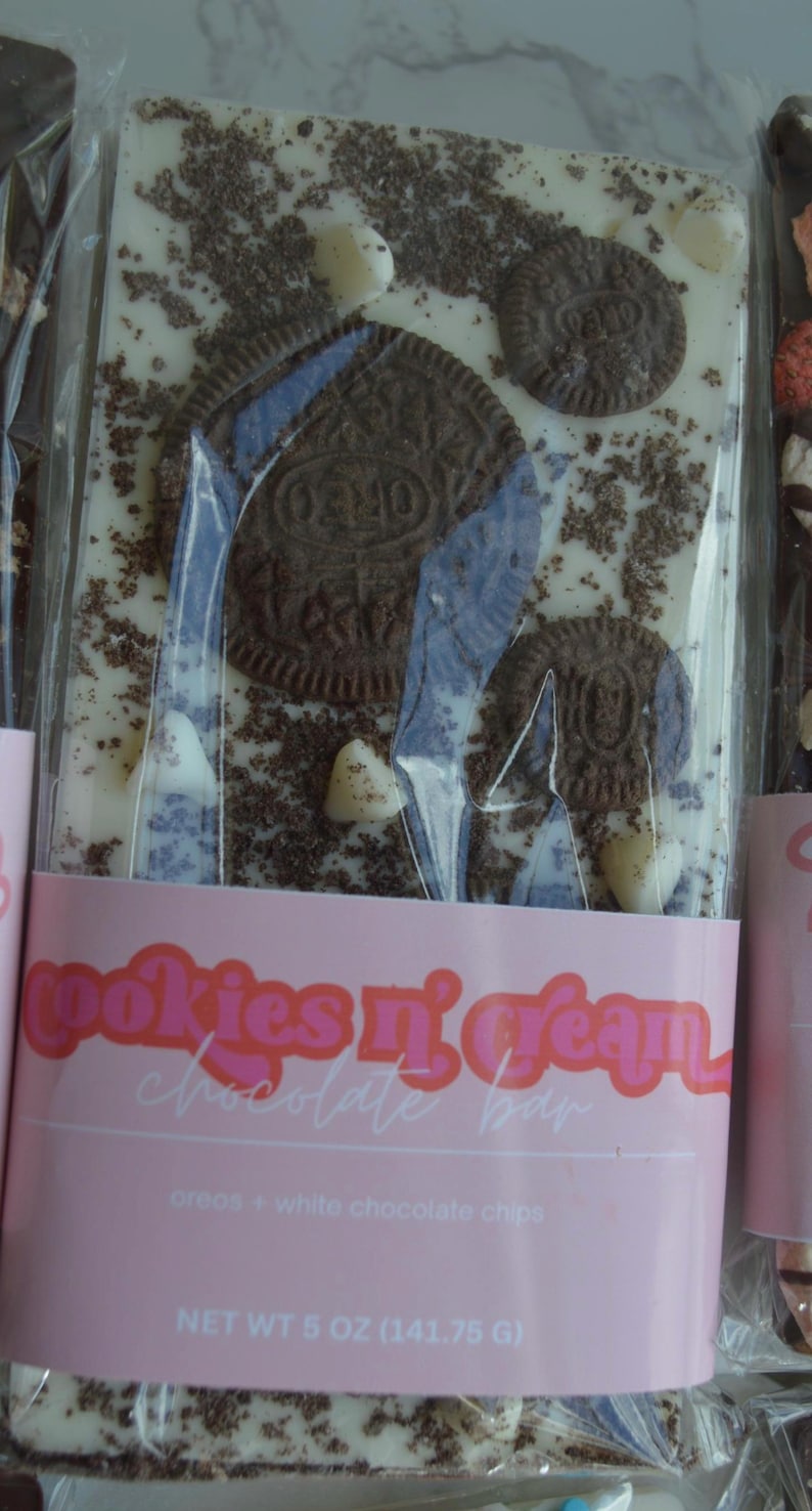 Peut inclure: Une barre de chocolat Cookies n' Cream avec des biscuits Oreo et des p&eacute;pites de chocolat blanc. La barre est envelopp&eacute;e dans du plastique transparent et porte une &eacute;tiquette rose avec le texte "Cookies n' Cream Chocolate Bar". Poids net 141,75 g.