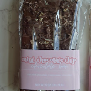 Peut inclure: Une barre de chocolat noir aux p&eacute;pites de menthe, emball&eacute;e dans du plastique transparent. L'&eacute;tiquette rose indique "mint chocolate chip chocolate bar" et "dark mint chocolate + semi-sweet chocolate". Poids net 141,75 g.