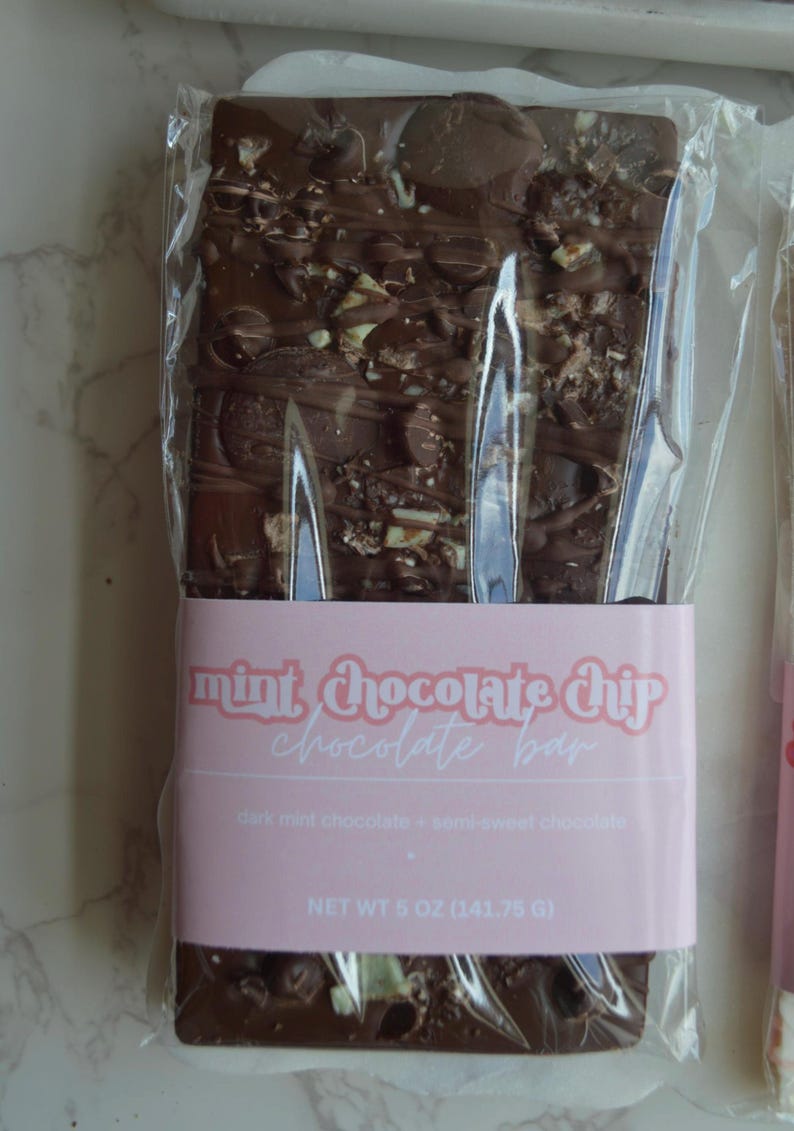 Peut inclure: Une barre de chocolat noir aux p&eacute;pites de menthe, emball&eacute;e dans du plastique transparent. L'&eacute;tiquette rose indique "mint chocolate chip chocolate bar" et "dark mint chocolate + semi-sweet chocolate". Poids net 141,75 g.