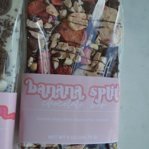 Peut inclure: Une barre de chocolat avec des chips de banane, des fraises s&eacute;ch&eacute;es et des cacahu&egrave;tes. La barre est emball&eacute;e dans du plastique transparent avec une &eacute;tiquette rose indiquant "Banana Split Chocolate Bar". Poids net 141,75 g.